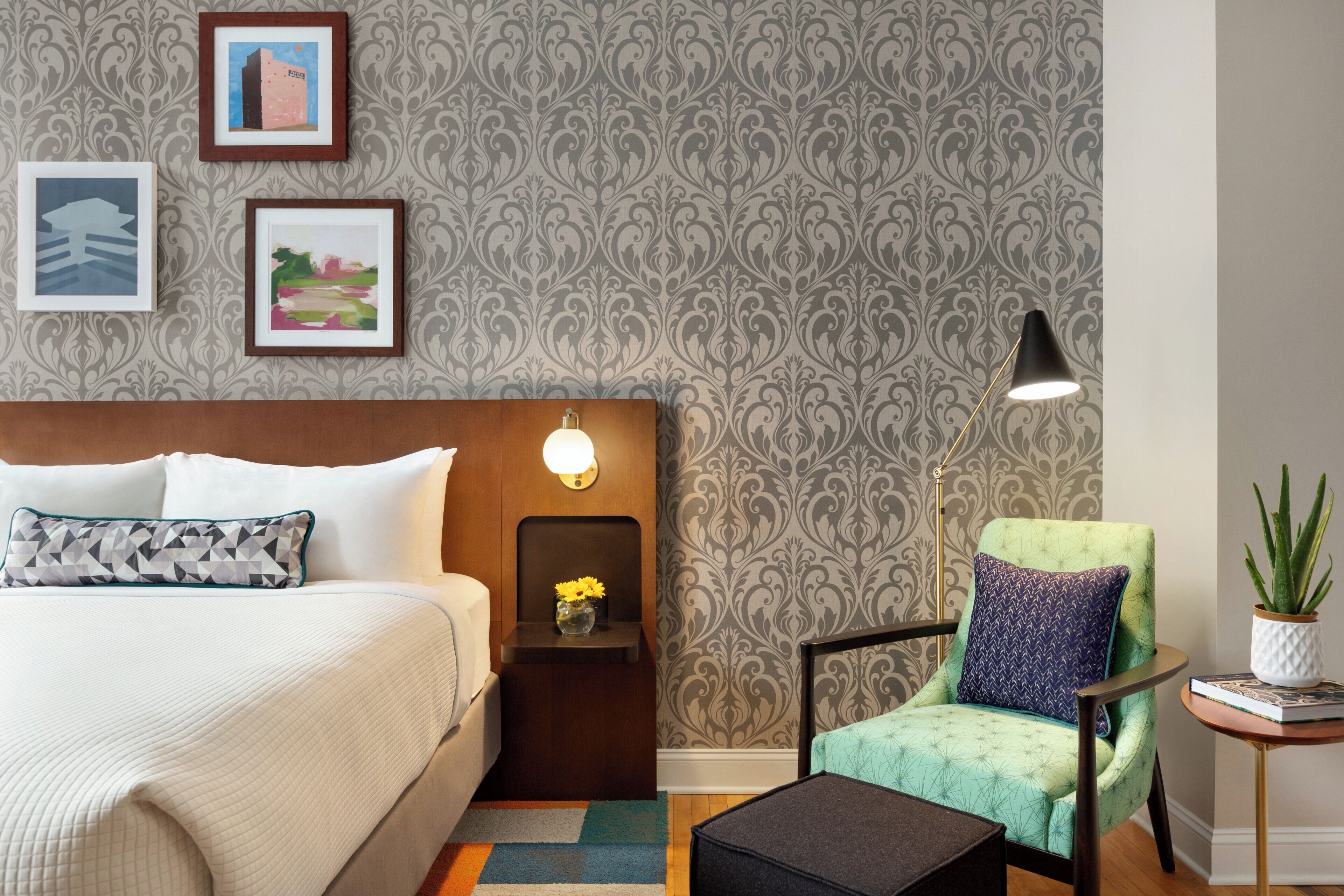 Une chambre d’hôtel avec un lit, une table de chevet avec une lampe, un fauteuil vert, une petite table avec une plante et des œuvres d’art encadrées accrochées au mur.