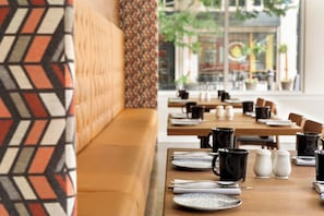 A restaurant interior with a long wooden table set for dining, a patterned wall, and a view of the street outside.