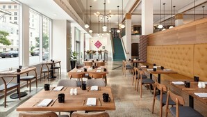 Un interior de restaurante moderno con mesas y sillas de madera, una escalera y amplios ventanales con vistas a la calle.