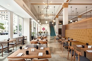 Un interior de restaurante moderno con mesas y sillas de madera, una escalera y amplios ventanales con vistas a la calle.