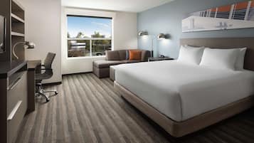 Quarto de hotel com uma cama grande, uma escrivaninha com cadeira, um sofá, uma mesa de cabeceira com abajur e vista para o exterior através de uma janela.