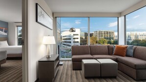 Una habitación de hotel moderna con un ventanal grande, un sofá, una mesita de noche con lámpara y una cama.