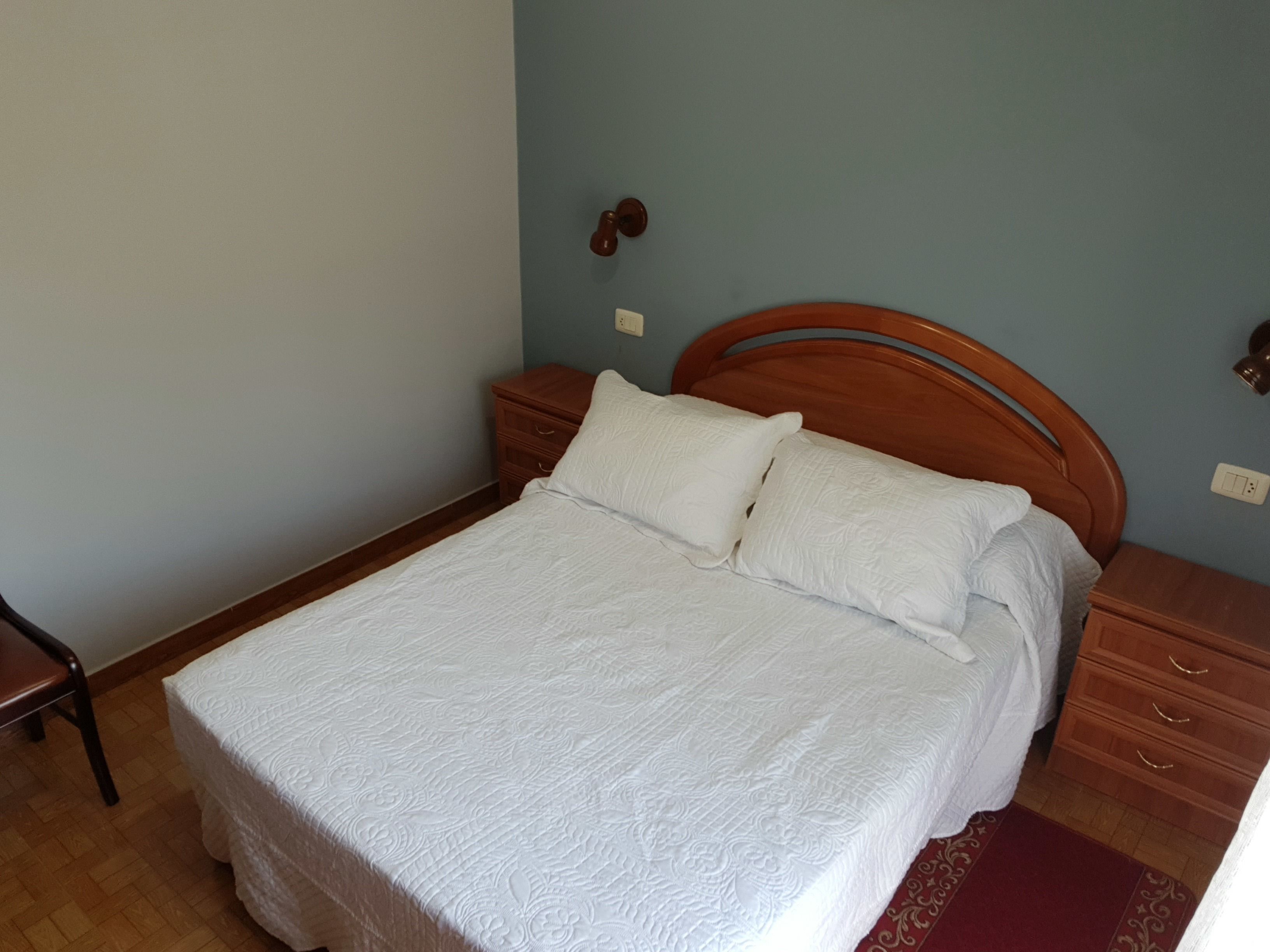 A neatly made bed with a wooden headboard and two pillows, a wooden nightstand with a lamp, and a wooden dresser with drawers.