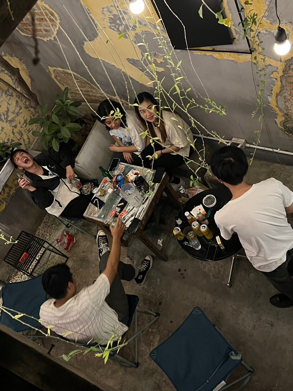 A group of people seated around a table with various items, in a room with a textured wall and hanging plants.