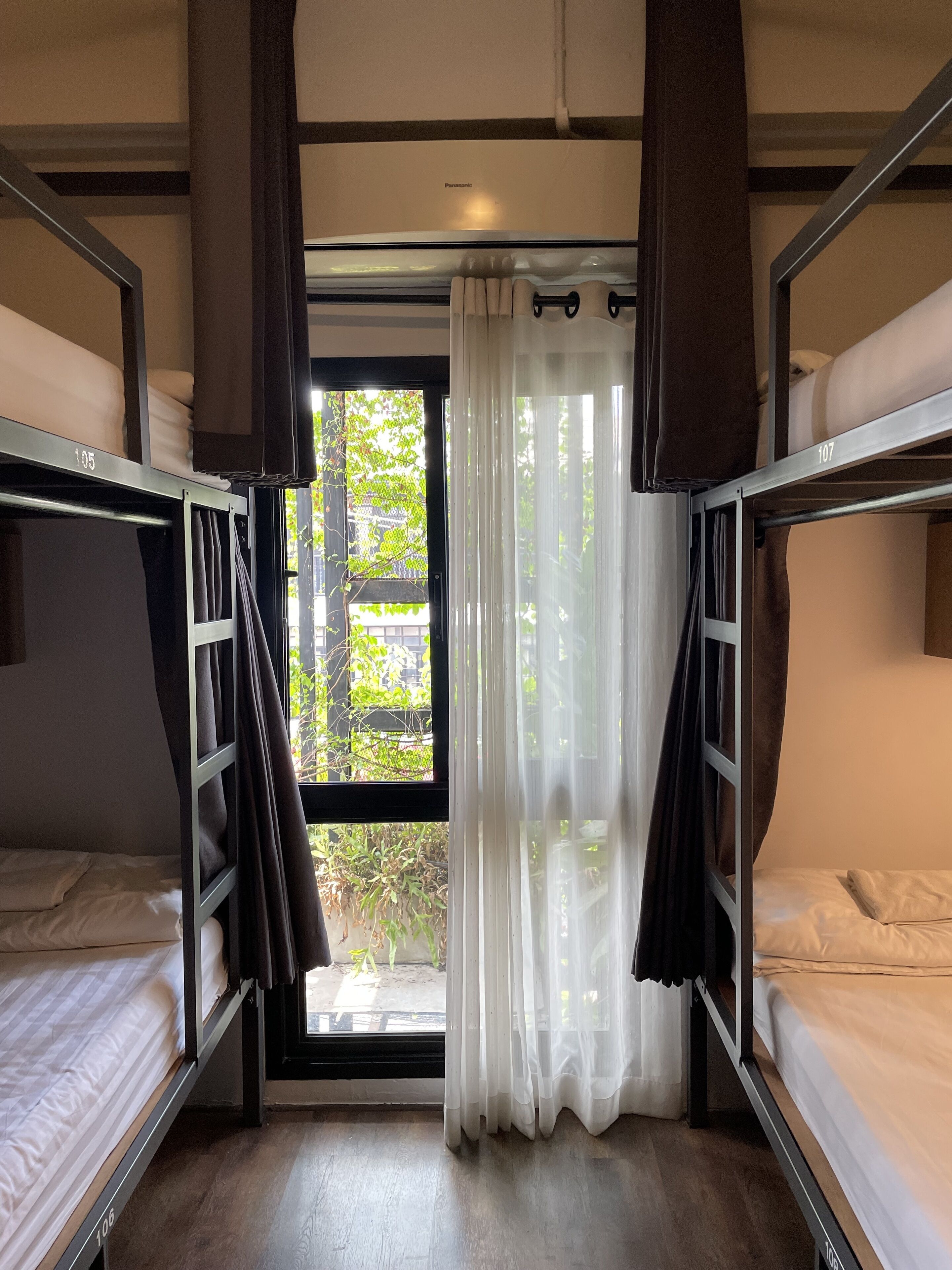 A dormitory room with bunk beds, a window with sheer curtains, and a view of greenery outside.