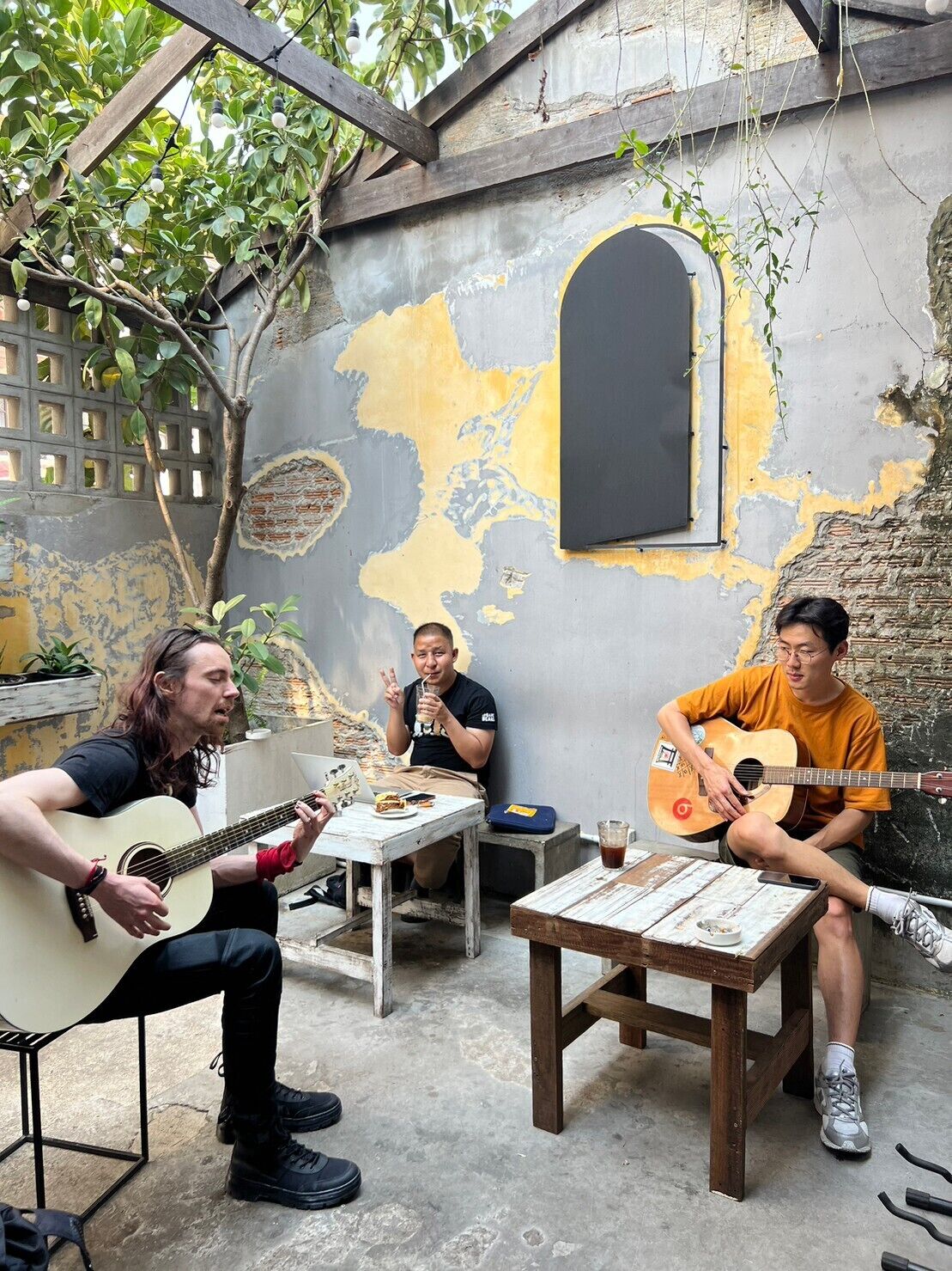 Three people playing guitars in a rustic outdoor setting with a weathered wall and a small table.