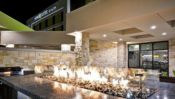 Exterior moderno de un hotel con fachada de piedra, una estructura con fuego en el centro y un cartel que dice 'Home Suites by Hilton'.