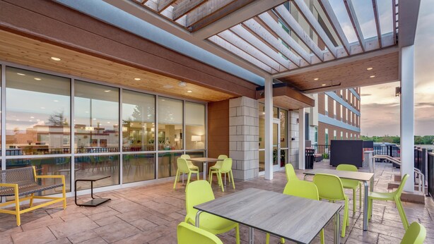 An outdoor patio with yellow and green chairs, a wooden table, and a view of the building's interior. - Home2 Suites by Hilton Indianapolis Northwest (Indianapolis)