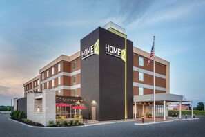 A modern hotel with 'HOME 2' signage, a flag, and a covered entrance. - Home2 Suites by Hilton Indianapolis Northwest (Indianapolis)