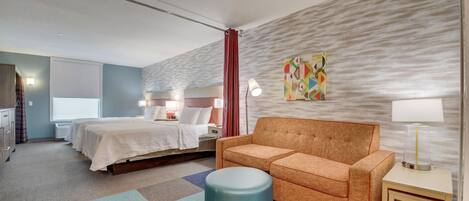 A hotel room with a bed, a sofa, a small table, a lamp, and a colorful artwork on the wall.