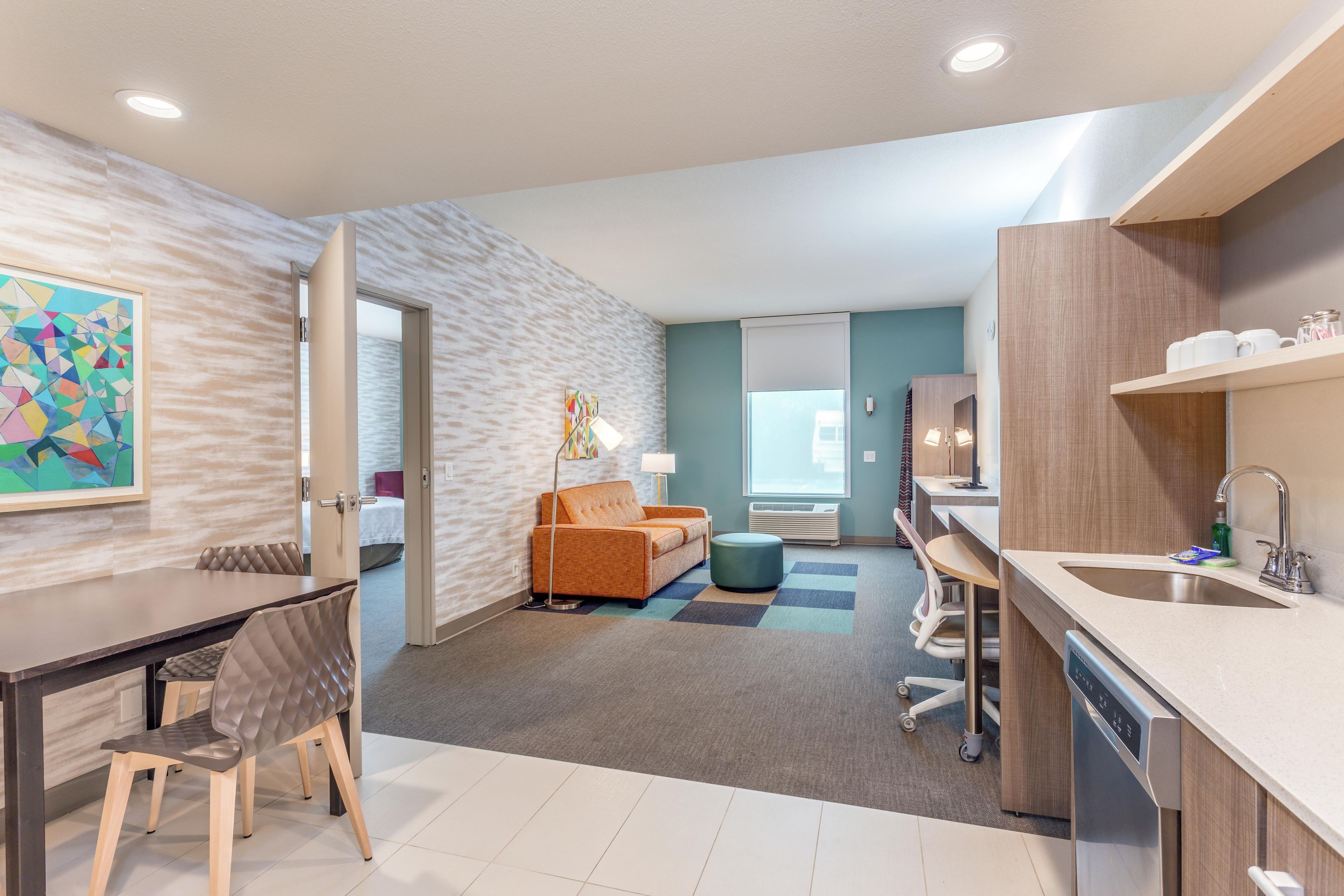 A modern hotel room with a dining area, kitchenette, and a view of the city.