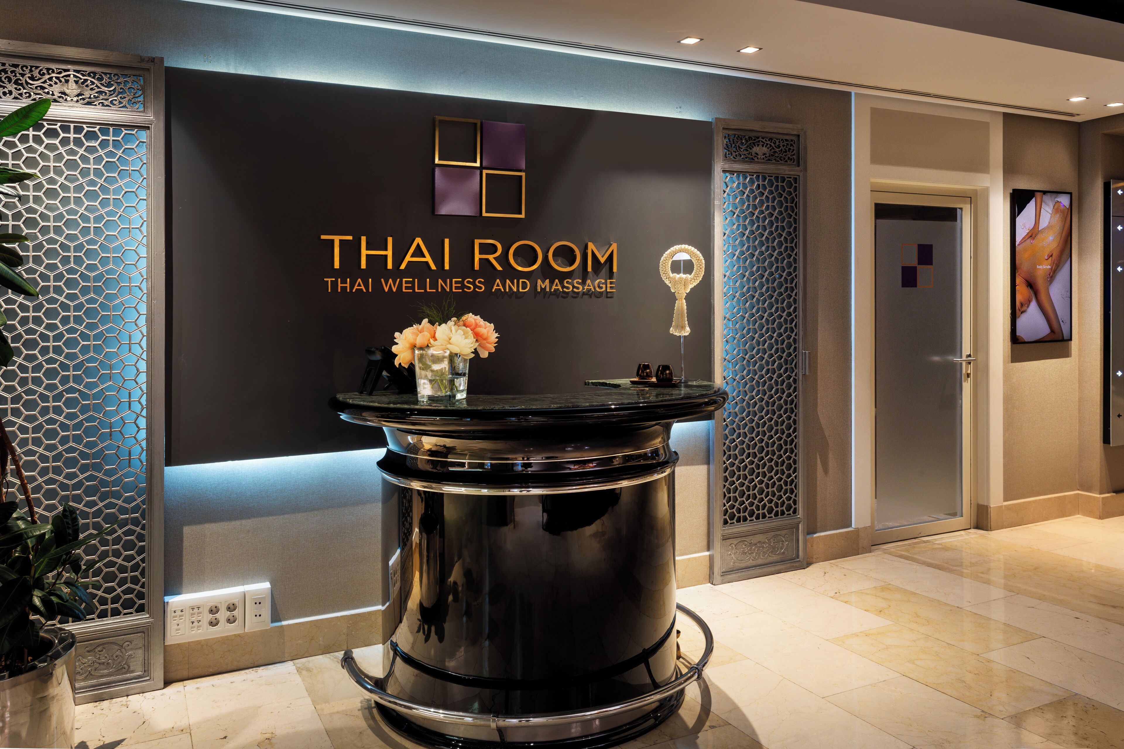 Zona de recepción con mostrador negro, un cartel en la pared que dice "Thai Room Thai Wellness and Massage", y un jarrón decorativo con flores.