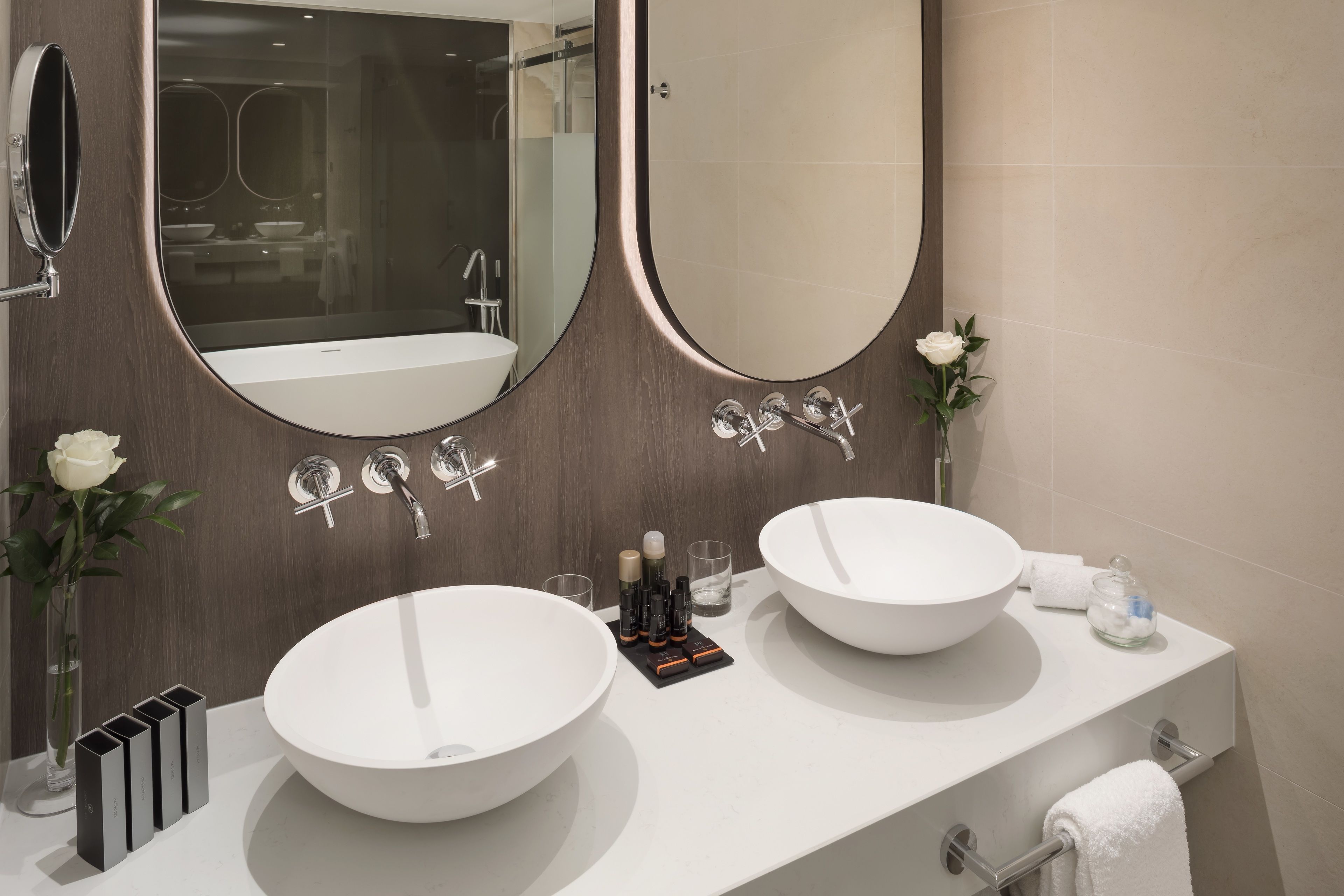 Un baño moderno con dos lavamanos grandes blancos, un espejo amplio y un florero con una rosa blanca.