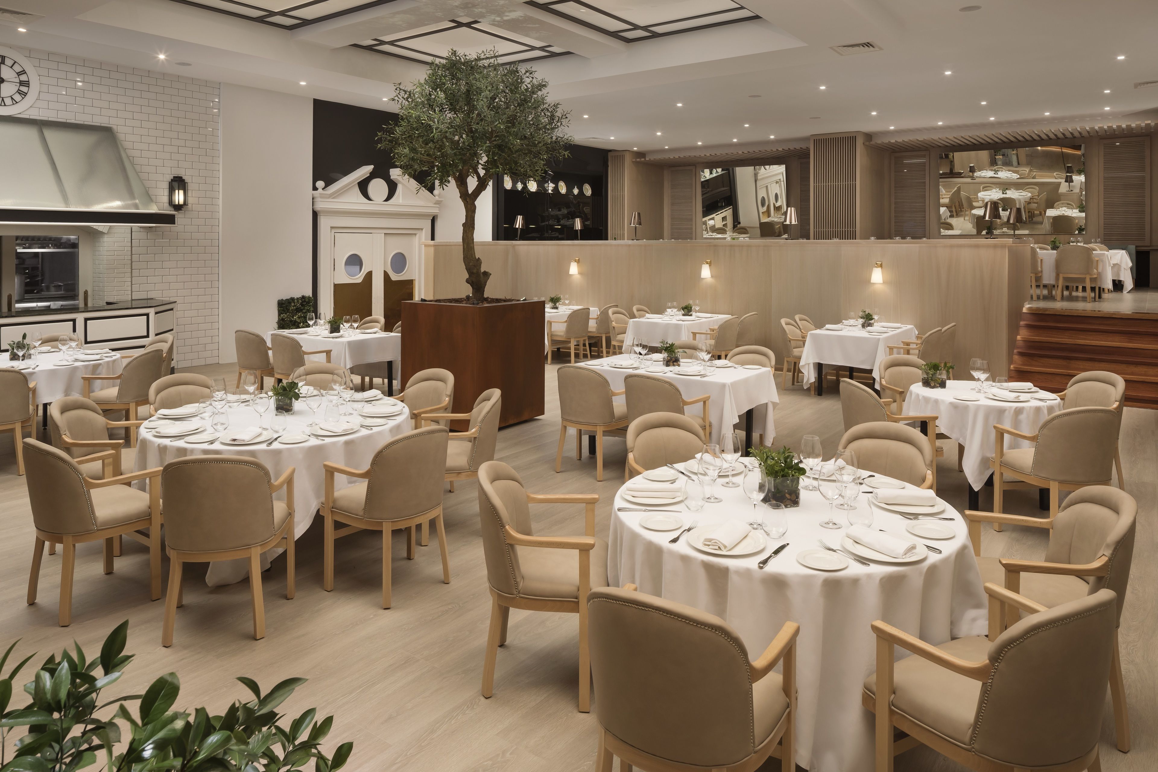 Un restaurante con mesas redondas dispuestas para comer, sillas color beige y un árbol en el centro.