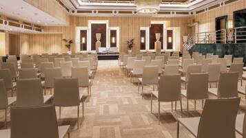 A conference room with rows of chairs and tables, a stage at the front, and a staircase to the right.