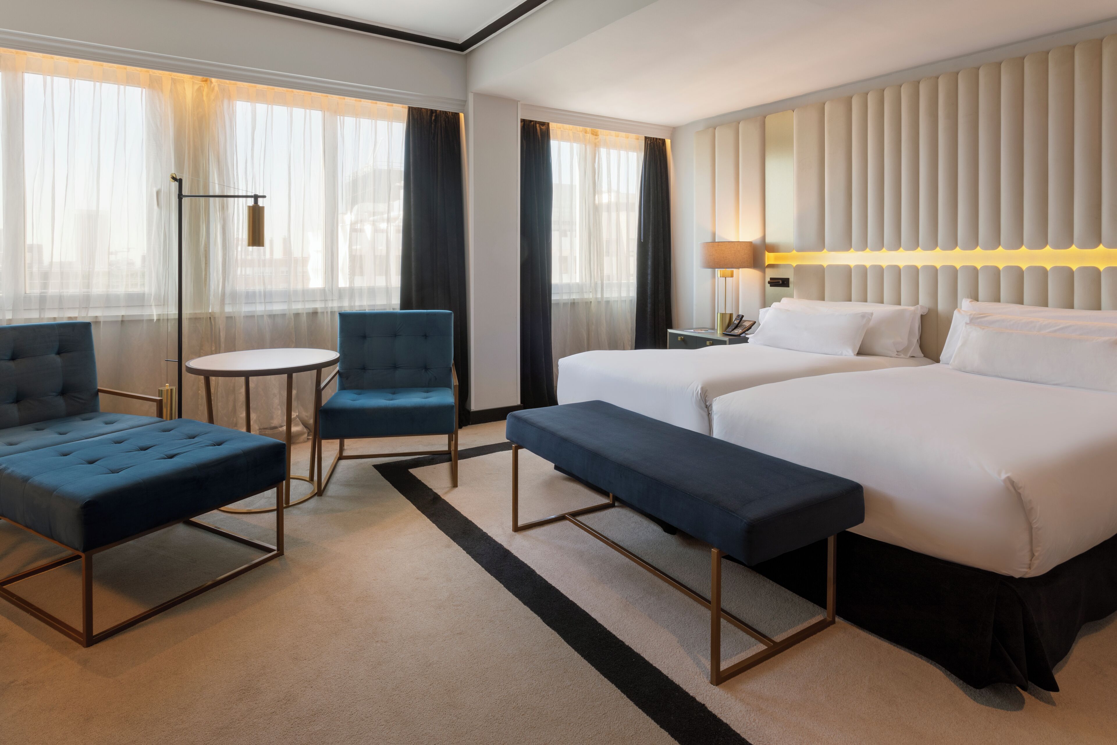 A hotel room with a large bed, two armchairs, a small round table, and a bench.