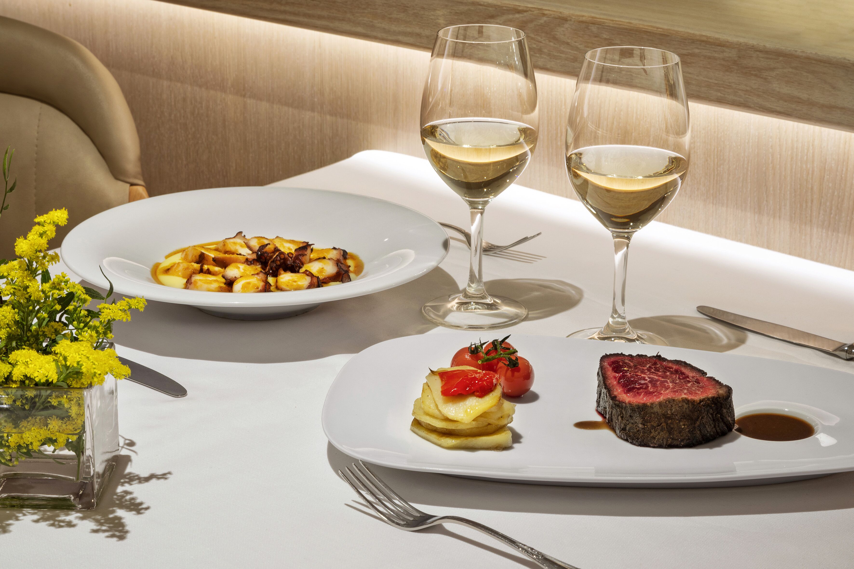 Una mesa de comedor con platos de comida, dos copas de vino y un florero con flores amarillas.