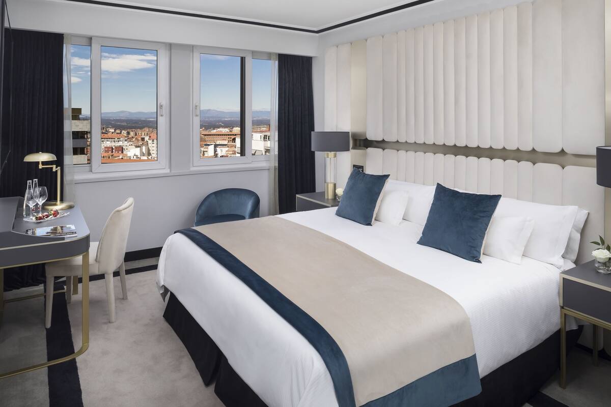 Ein Hotelzimmer mit einem großen Bett, einem Schreibtisch, einem Stuhl und Blick auf die Stadt durch große Fenster.