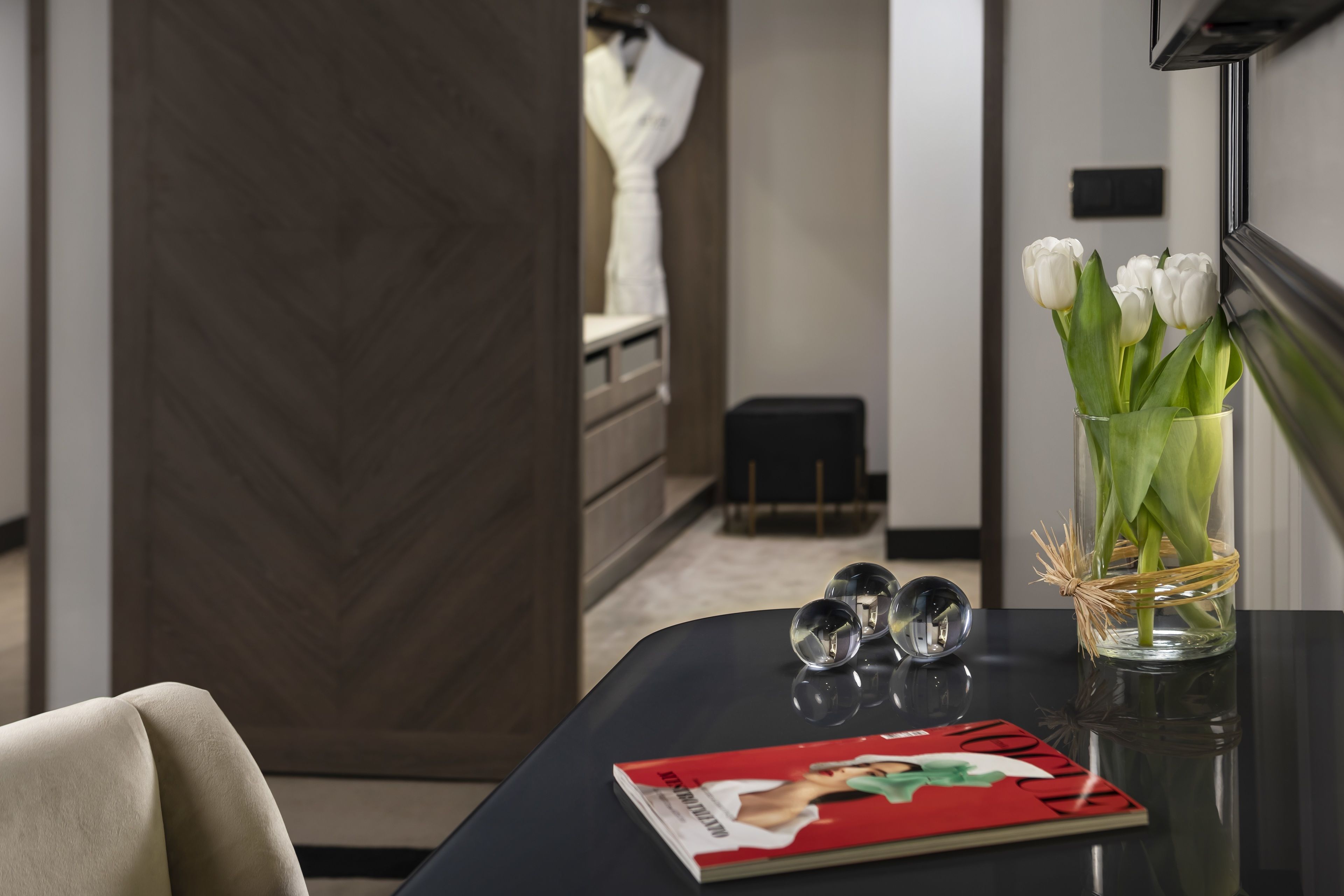 Quarto de hotel moderno com um guarda-roupa de madeira escura, um vaso de vidro com flores brancas, uma revista e um pequeno alto-falante preto sobre uma mesa preta.