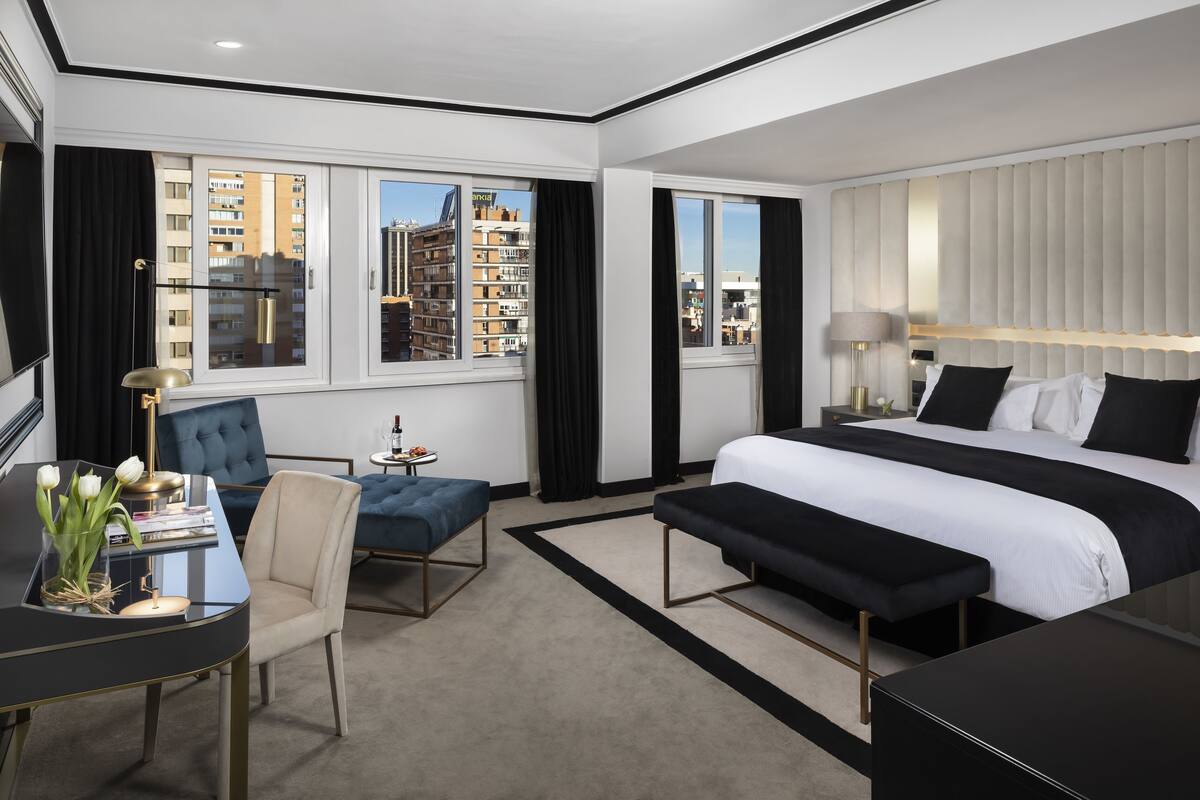 Ein modernes Hotelzimmer mit einem großen Bett, einem Schreibtisch mit Lampe, einem Sessel und Blick auf die Stadt durch große Fenster.