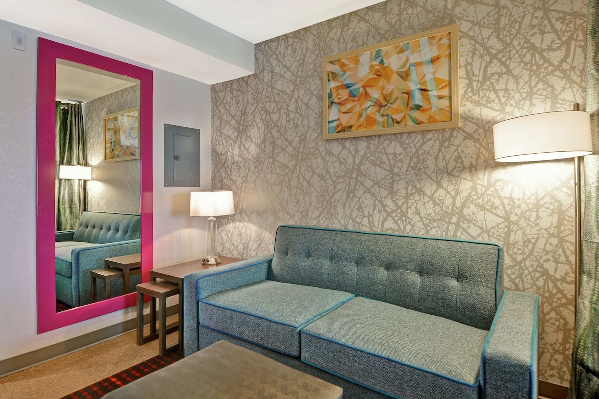 Una habitación de hotel moderna con un sofá modular, una mesita, una lámpara y un espejo que refleja un rincón de descanso acogedor.