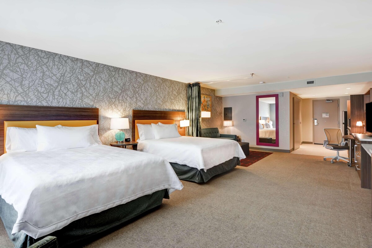 Een hotelkamer met twee bedden, een bureau, een stoel en een badkamer zichtbaar door een geopende deur.