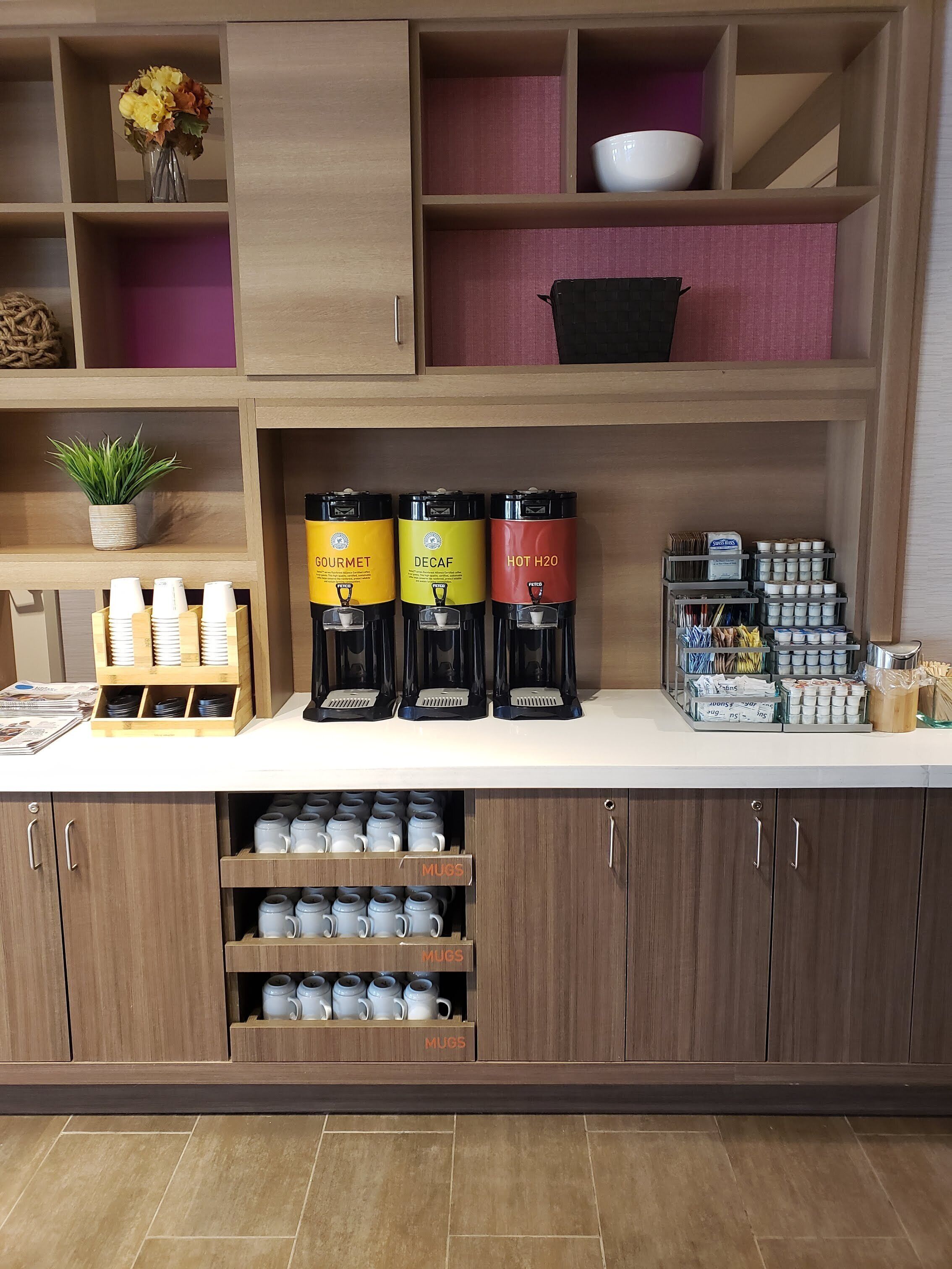 Un espace dédié au café, équipé de trois distributeurs de café étiquetés « Gourmet », « Décaféiné » et « Eau chaude ». En dessous, on trouve des étagères avec des tasses et une petite plante en pot.