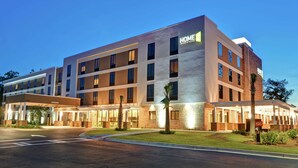 Un hôtel à plusieurs étages au design moderne, avec une enseigne « HOME2 » bien visible, un espace extérieur couvert et une pelouse bien entretenue.