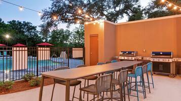 Une terrasse extérieure avec une longue table et des chaises, un barbecue et une piscine en arrière-plan.