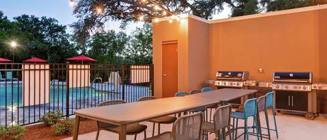 An outdoor patio with a long table and chairs, a barbecue grill, and a pool in the background.
