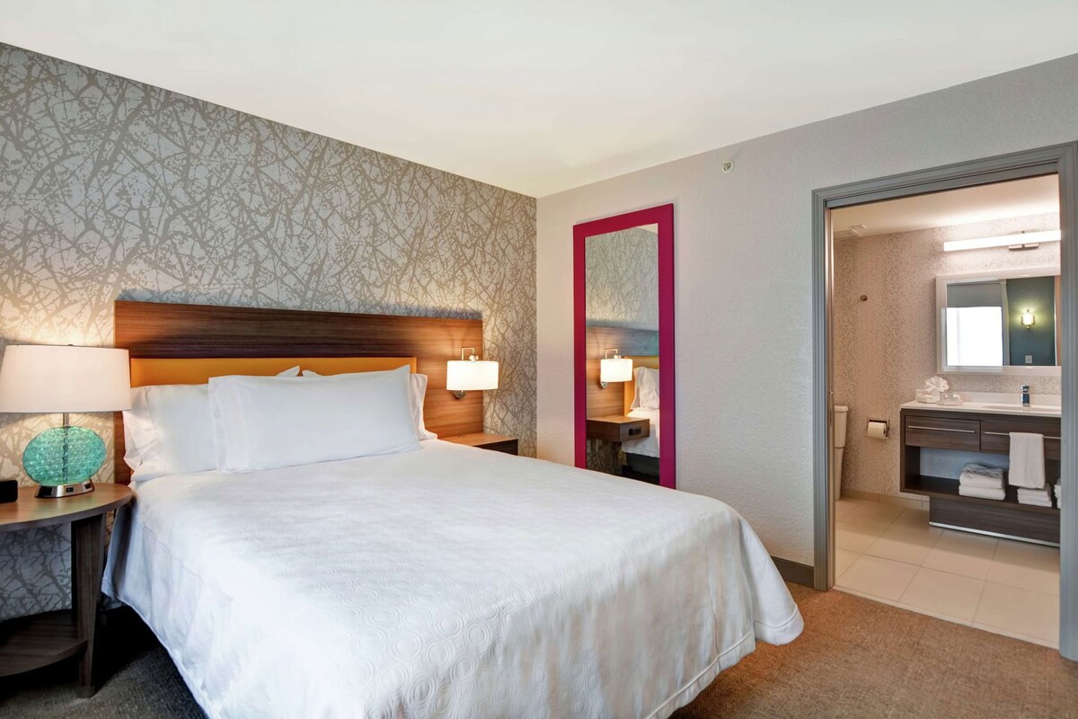 Hotelkamer met een bed, nachtkastje met lamp, spiegel en badkamer met wastafel en handdoekrek.