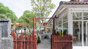 Uma entrada com portão e cerca de bambu, área de estar coberta ao ar livre e porta de vidro que dá para um edifício com telhado de telhas.