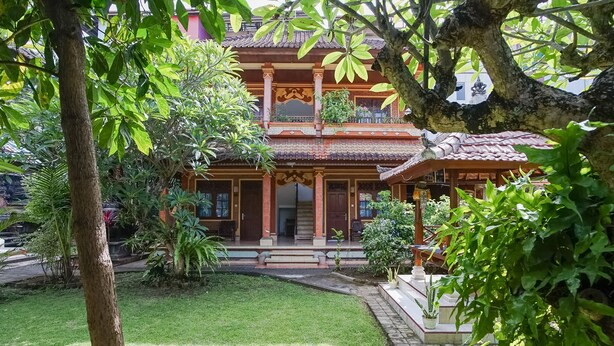 Um edifício tradicional com telhado de telhas, entrada central com degraus e um jardim circundante com diversas plantas.