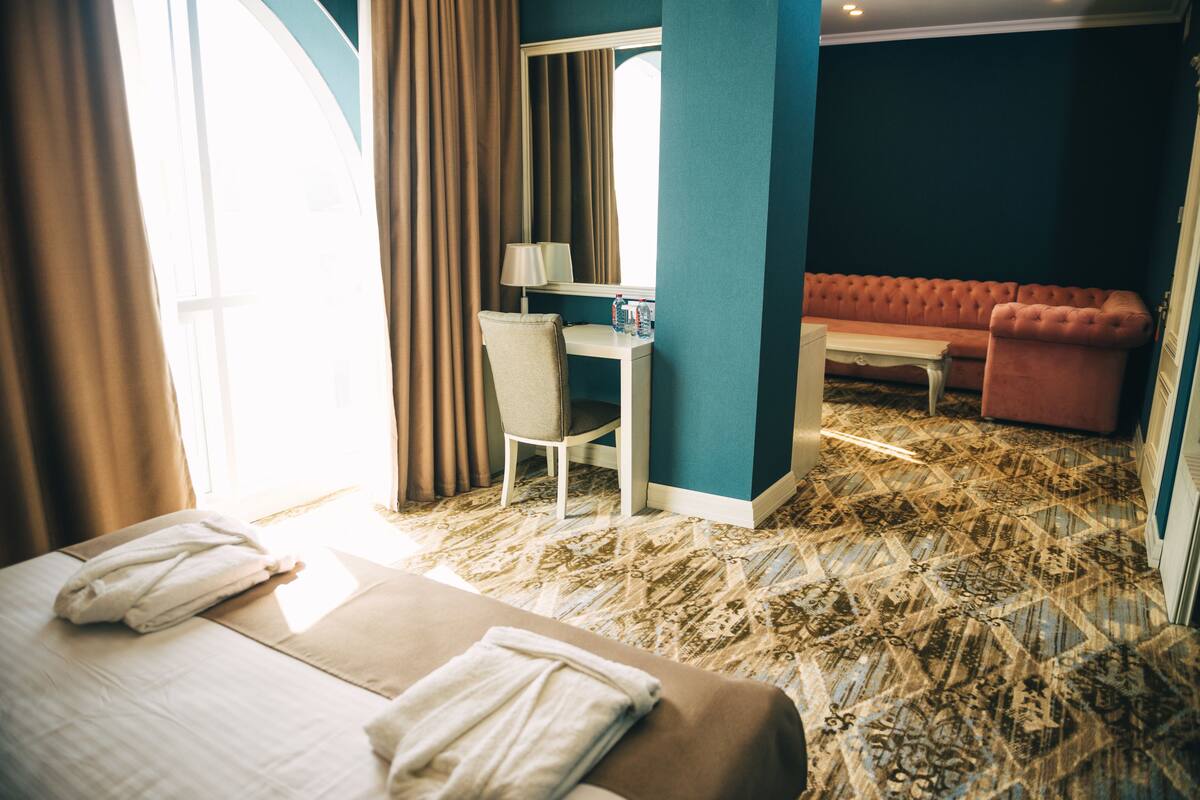 A hotel room with a bed, a desk, a chair, a sofa, and a large window with curtains.