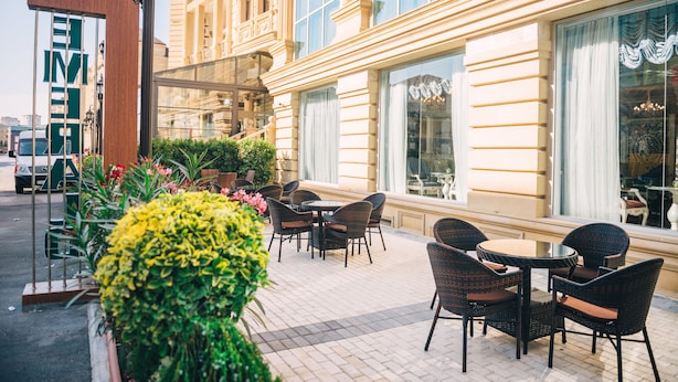 An outdoor seating area with wicker chairs and tables, a large window with curtains, and a building facade with large windows and decorative elements. - Emerald Suite Hotel (Baku)