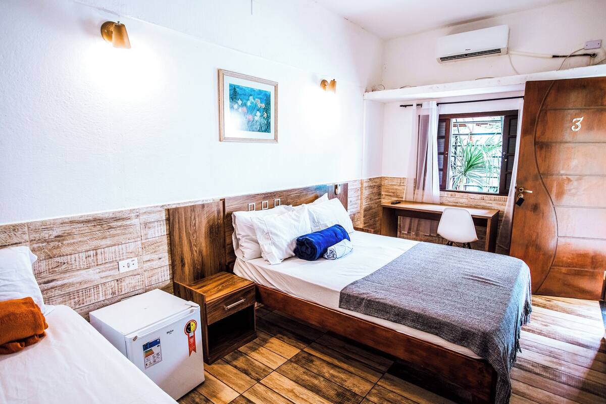 A hotel room with a wooden headboard bed, a small bedside table, a refrigerator, a window with a view of greenery, and a wooden door.