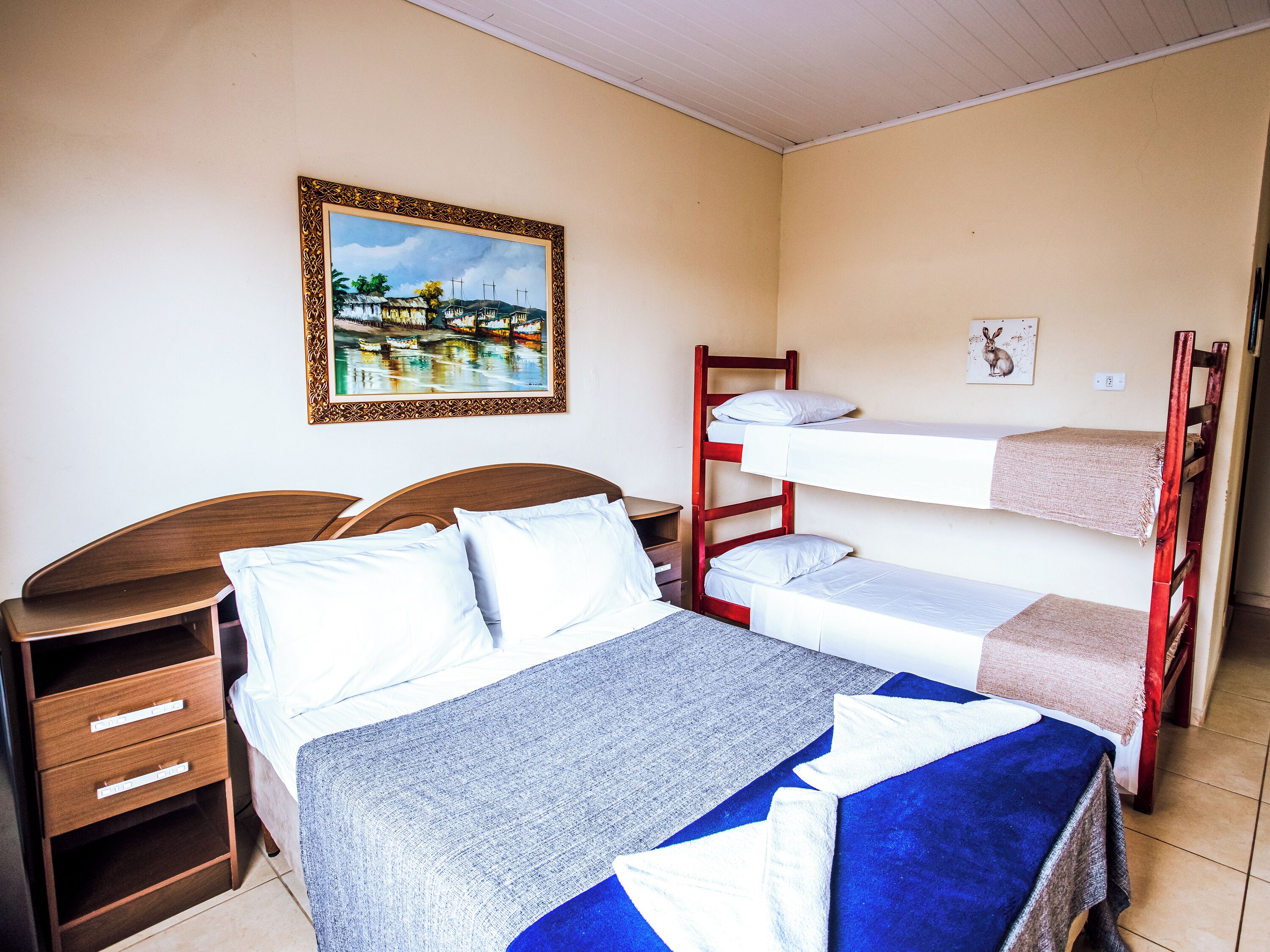 A room with a bunk bed, a single bed, a wooden nightstand, and a painting on the wall.