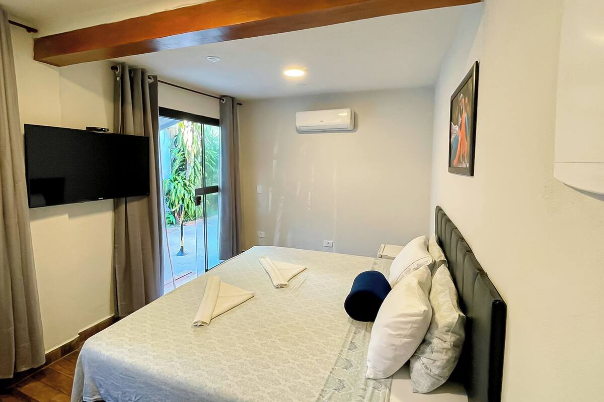 A hotel room with a bed, a flat-screen TV mounted on the wall, an air conditioning unit, and a door leading to a balcony with a view of greenery.