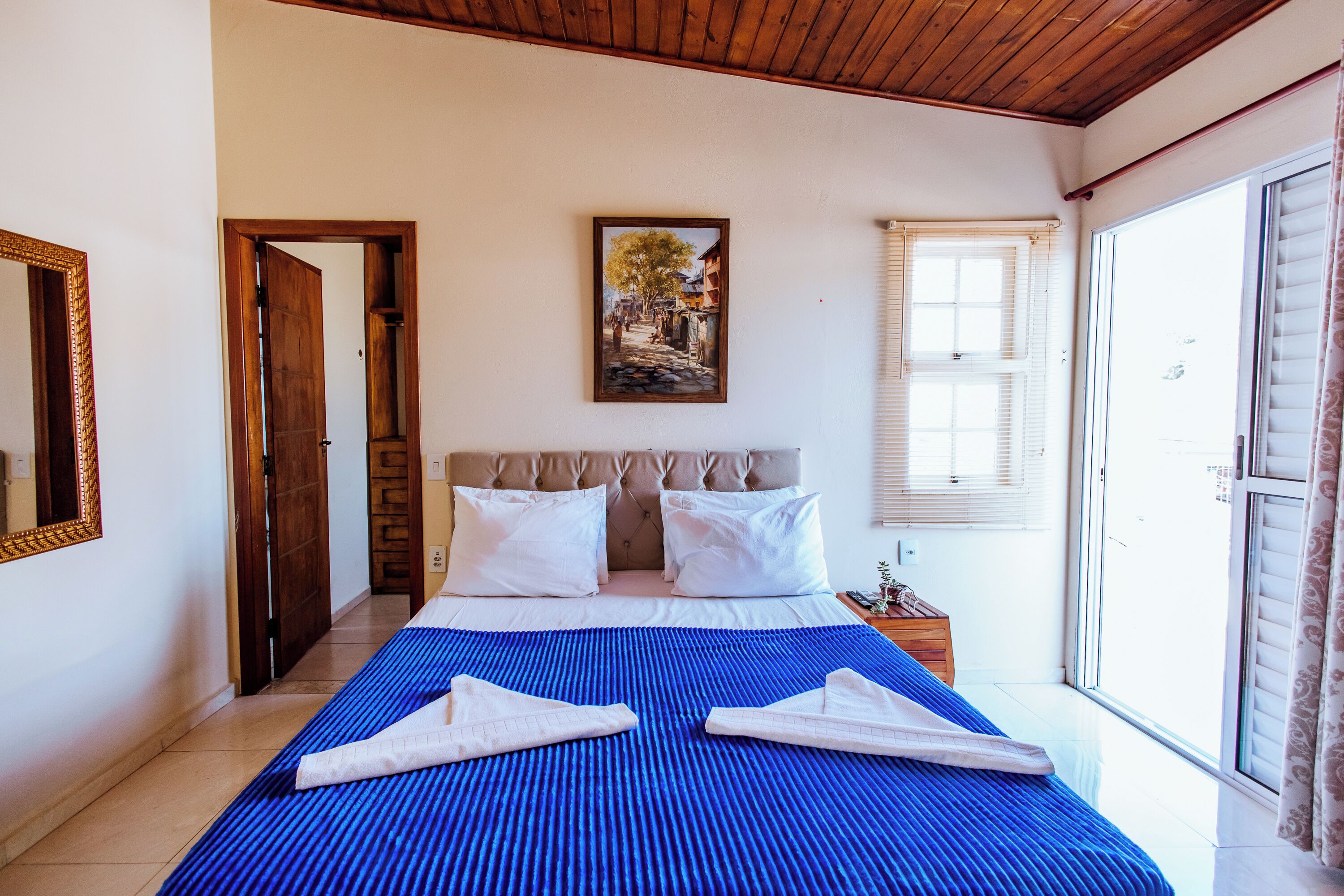A bedroom with a large bed, a painting on the wall, and a window with blinds.