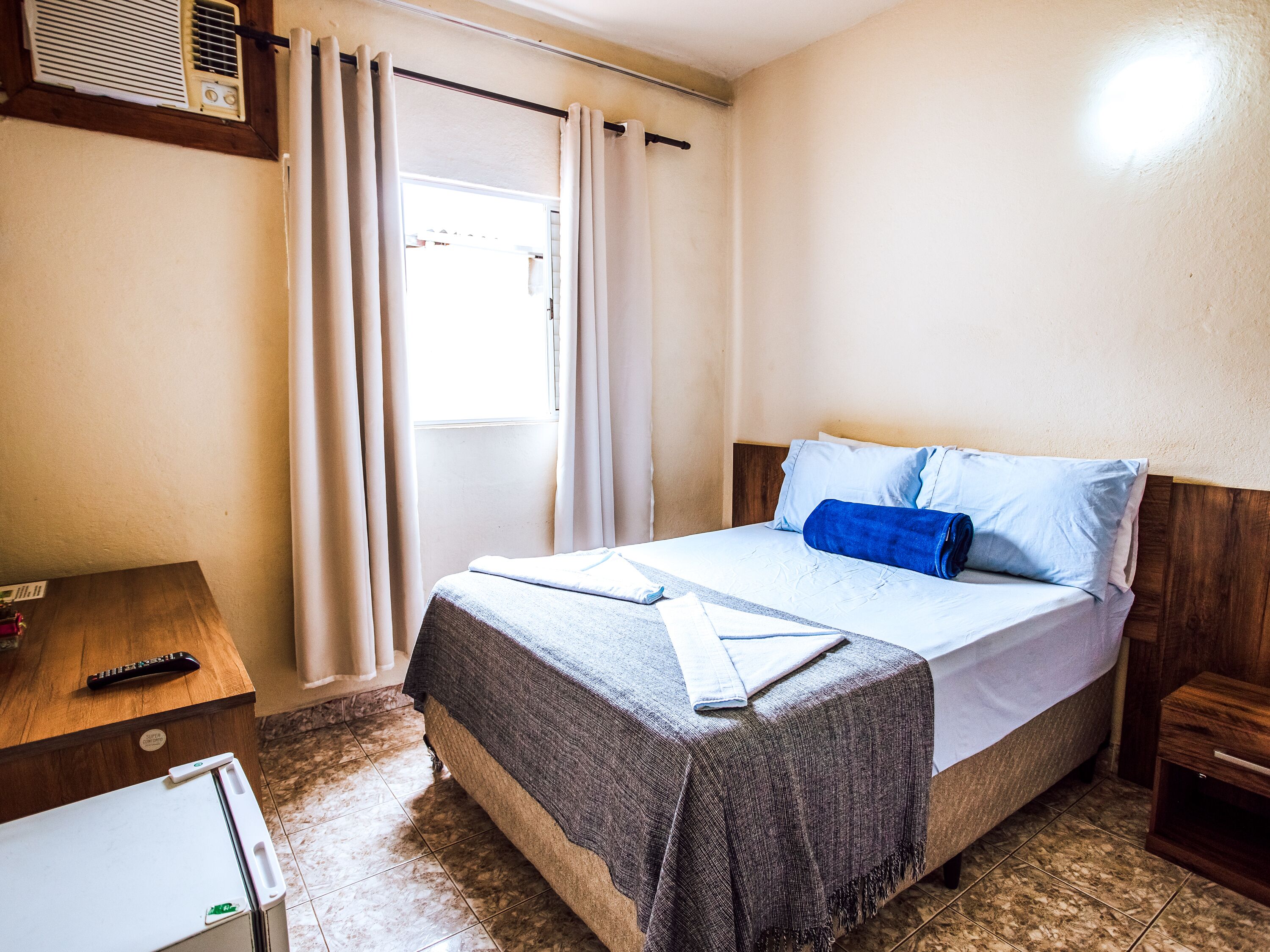 A hotel room with a bed, a wooden nightstand, a remote control, and a window with curtains.