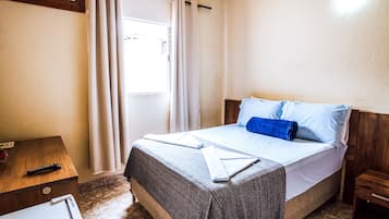 A hotel room with a bed, a wooden nightstand, a remote control, and a window with curtains.