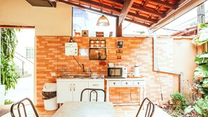 A compact kitchen with a brick wall, a microwave, a sink, and a small refrigerator. There is a dining table with chairs and a hanging light fixture above it. - VELINN Pousada Praia do Perequê (Ilhabela)