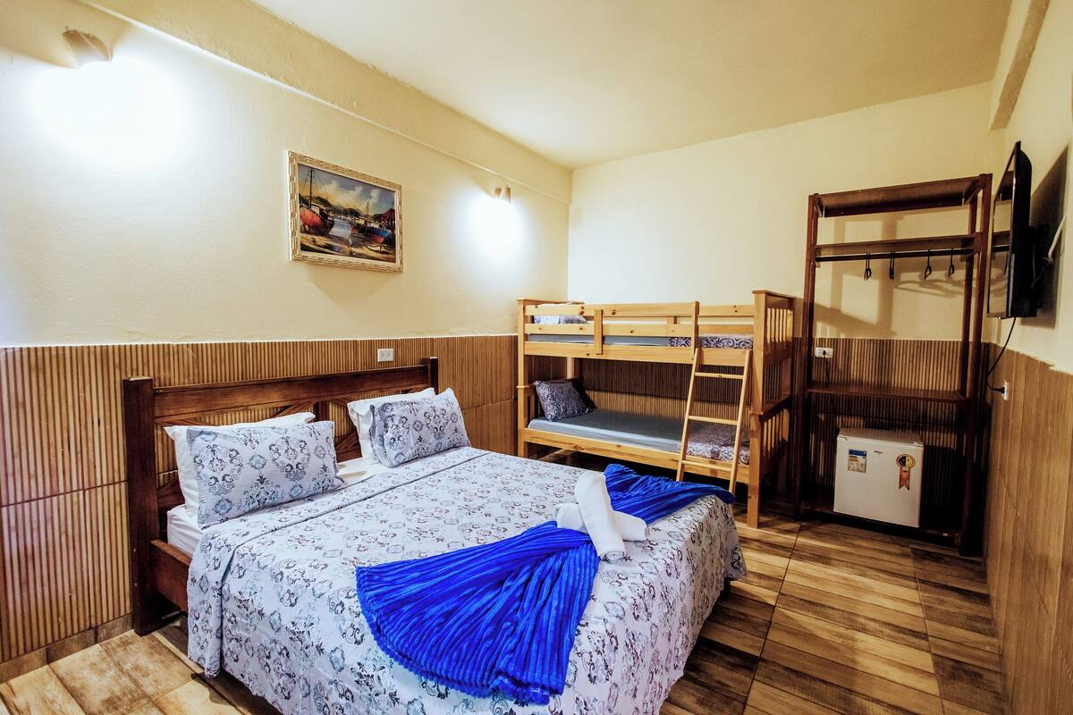 A bedroom with a wooden bunk bed, a single bed with patterned bedding, a small refrigerator, and a wall-mounted TV.