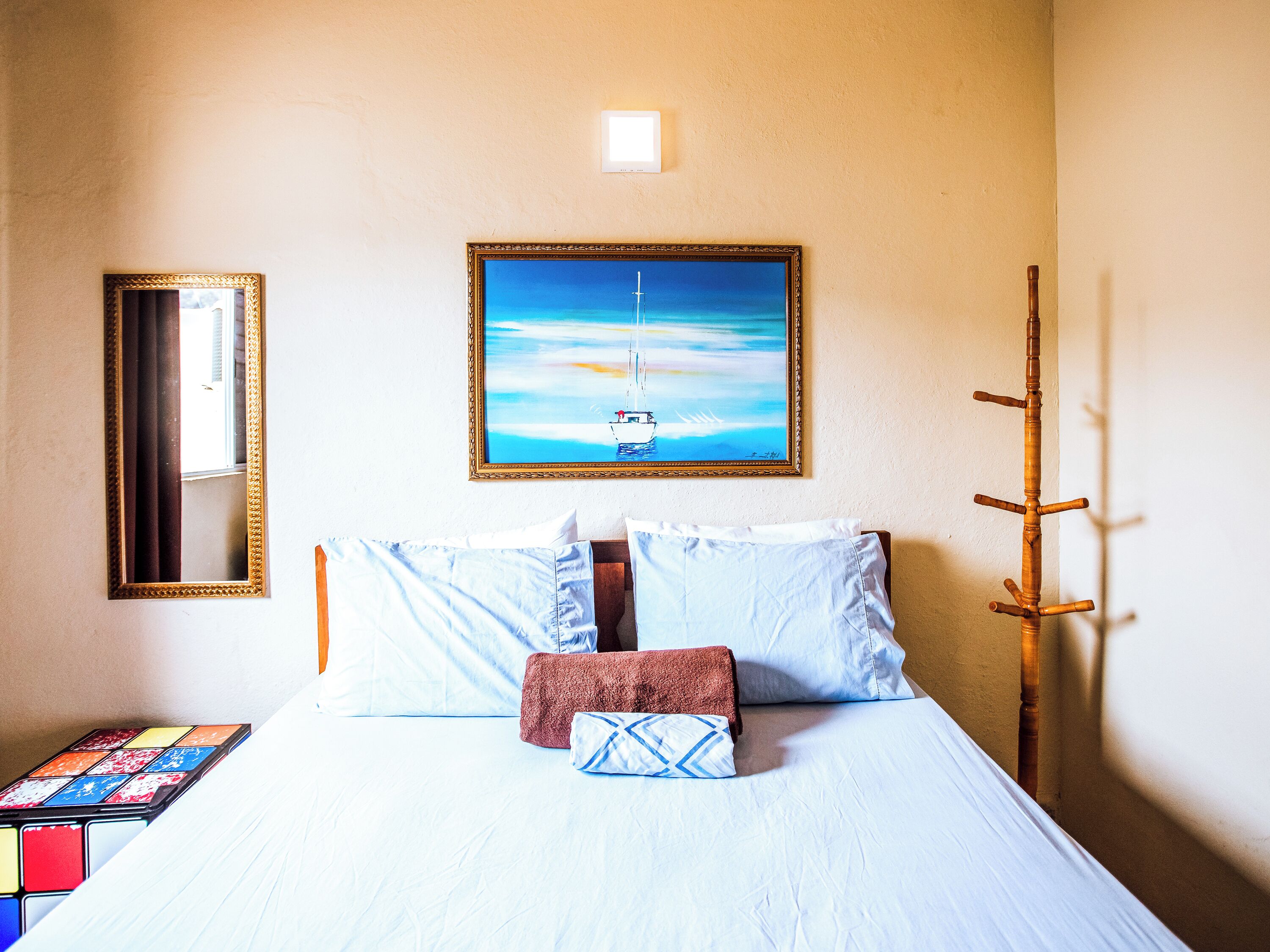 Habitación de hotel con cama, almohadas, un cuadro de un velero, un espejo y un perchero de madera.
