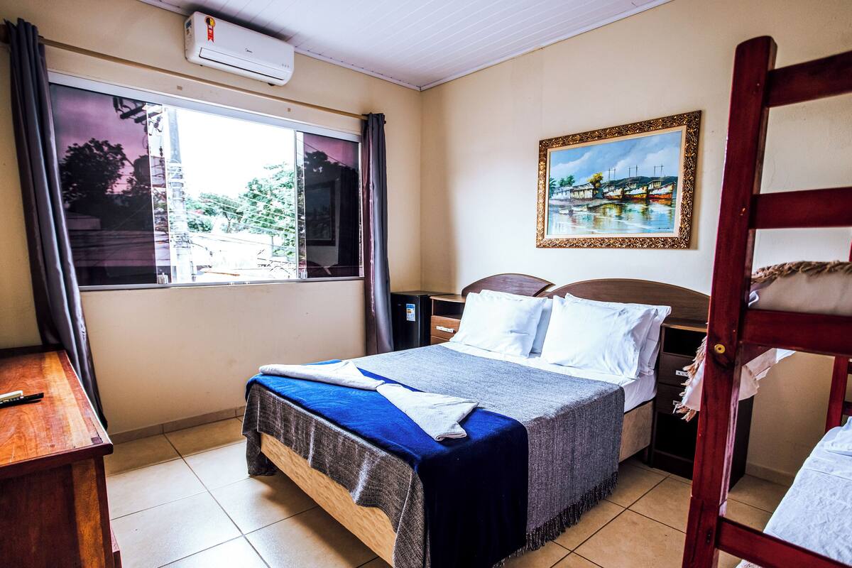 A hotel room with a bed, a nightstand, a desk, a window with curtains, a painting on the wall, and a wooden chair.