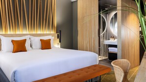 A modern hotel room with a large bed, a wooden headboard, a bench, a wicker chair, and a bathroom with a modern sink and mirror.