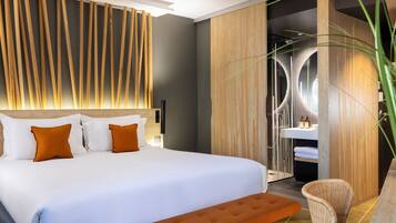 Une chambre d’hôtel moderne avec un grand lit, une tête de lit en bois, un banc, un fauteuil en osier et une salle de bain dotée d’un lavabo et d’un miroir contemporains.