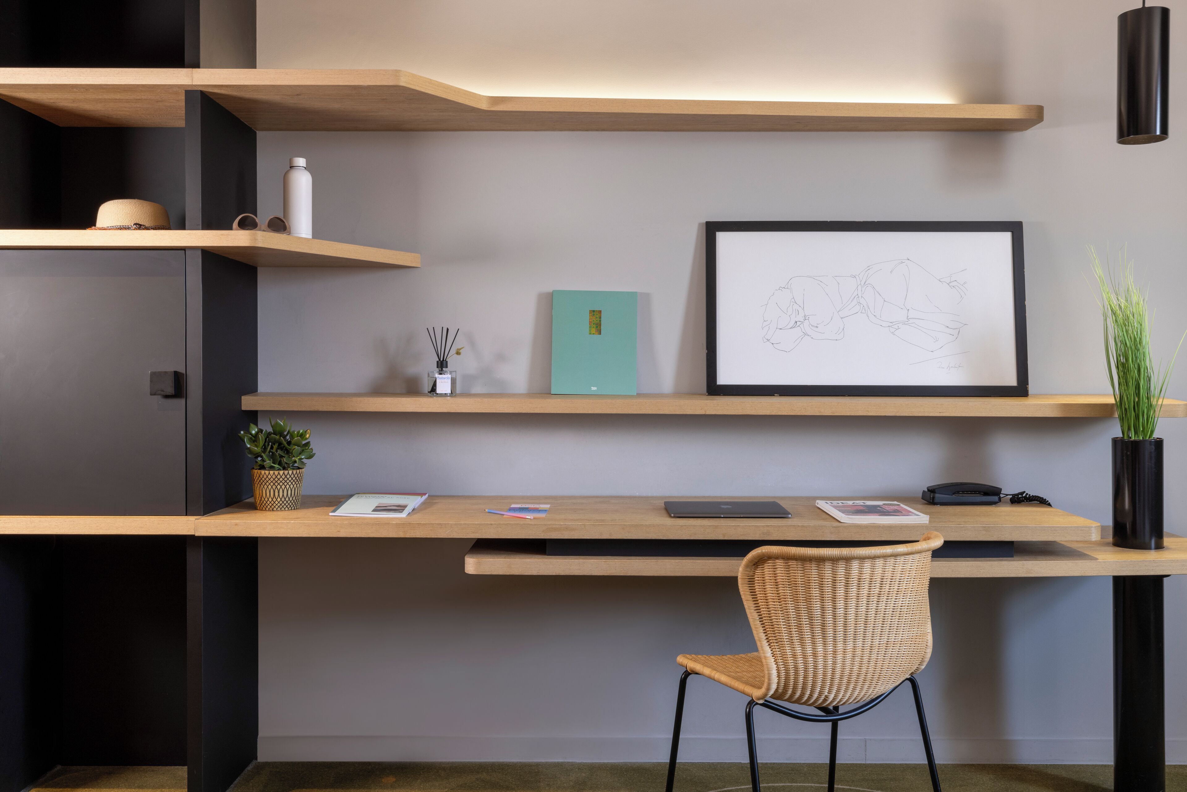 Een moderne werkplek met een houten bureau, een rieten stoel en planken met verschillende spullen, waaronder een plant, een waterfles en een ingelijste tekening.