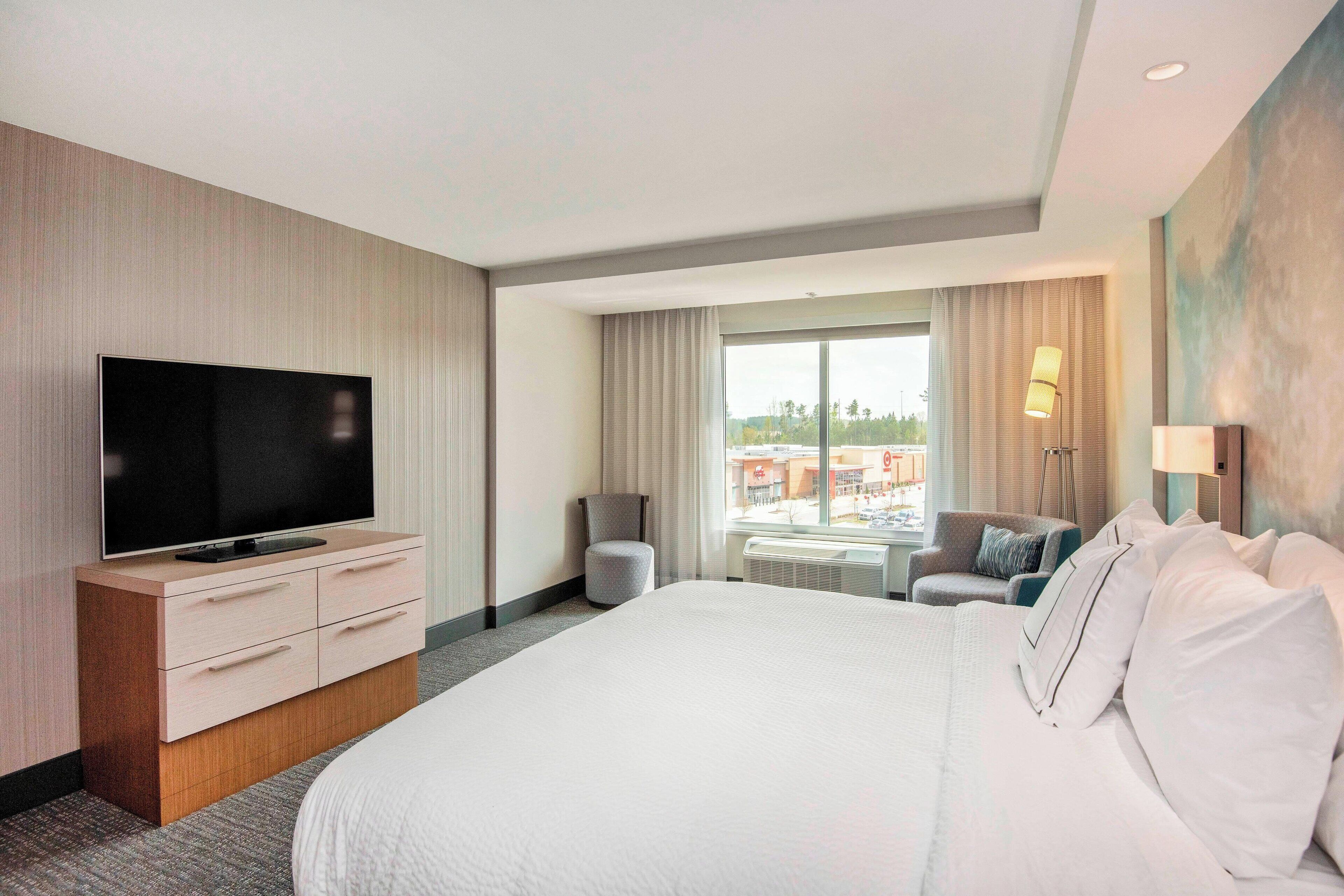 Une chambre d’hôtel avec un grand lit, un téléviseur à écran plat posé sur une commode en bois, un petit fauteuil, une lampe et une vue sur l’extérieur par une grande fenêtre.