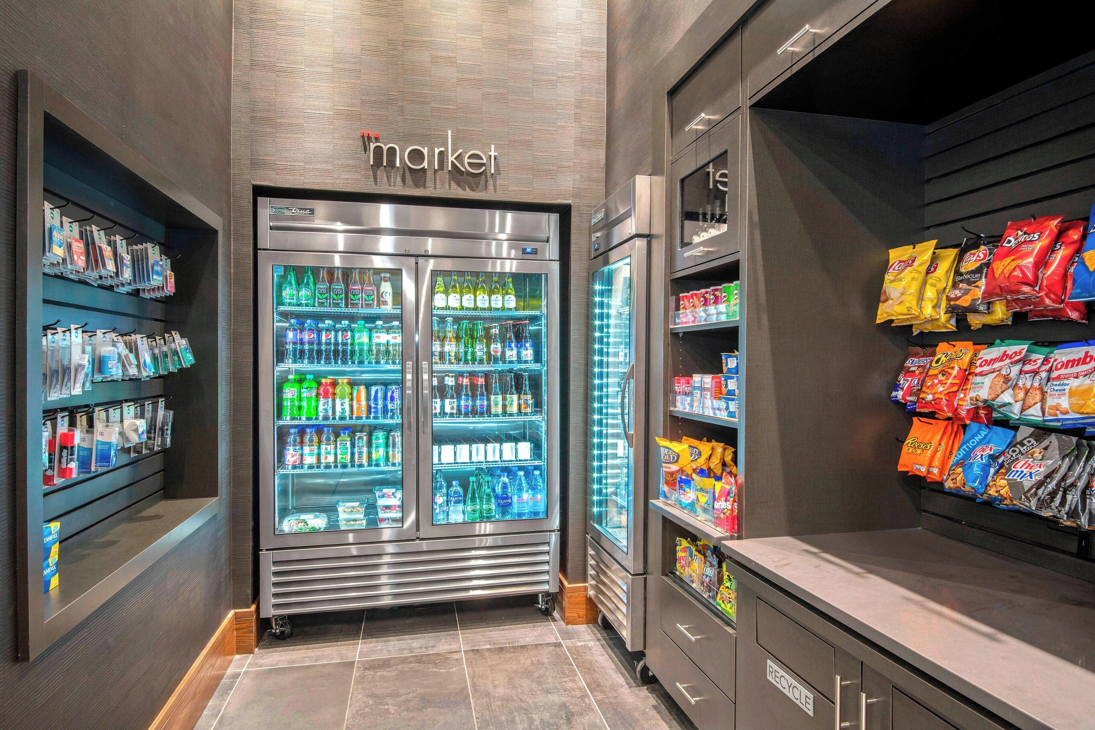 Mostrador refrigerado de bebidas con un cartel que dice 'market', rodeado de estantes con aperitivos y productos envasados.